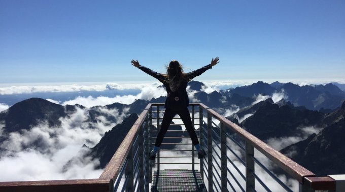 Person Standing On Hand Rails With Arms Wide Open Facing The Mountains And Clouds